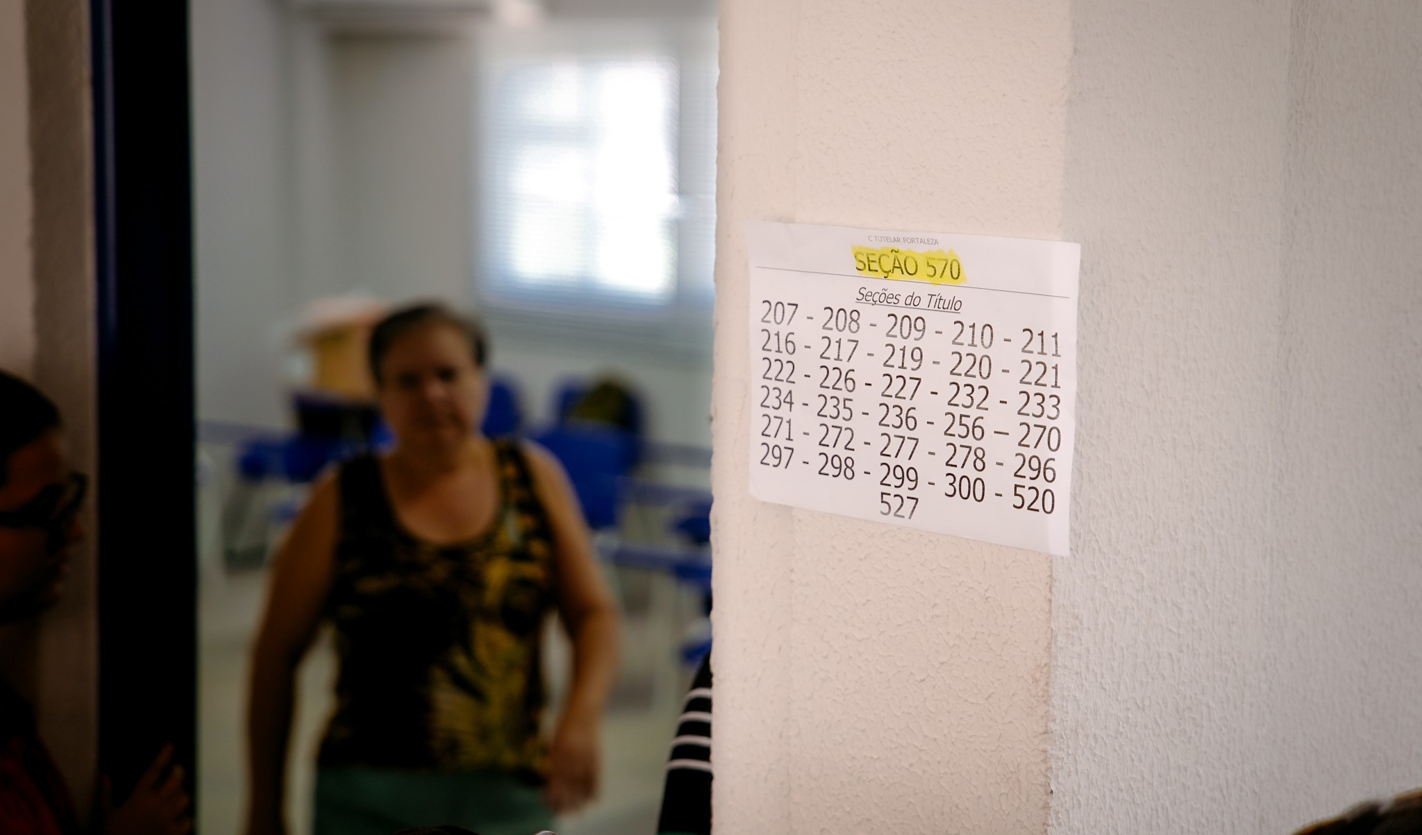 a foto mostra uma porta com um cartaz onde estão escritas as seções de votações. Ao fundo, uma mulher sai da sala